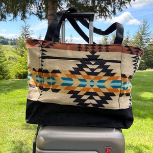May include: A large tote bag with a Southwestern geometric pattern in black, orange, and turquoise on a cream background. The bag has black handles and a black bottom. It is sitting on top of a grey suitcase with a retractable handle.