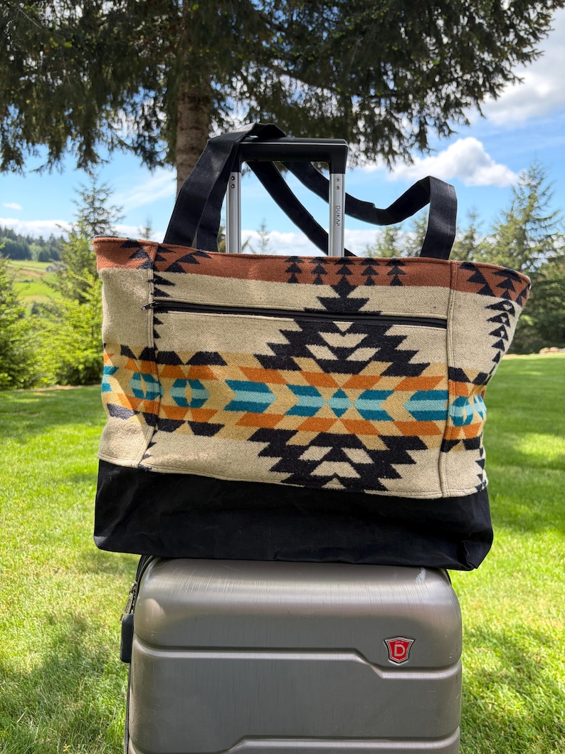 May include: A large tote bag with a Southwestern geometric pattern in black, orange, and turquoise on a cream background. The bag has black handles and a black bottom. It is sitting on top of a grey suitcase with a retractable handle.