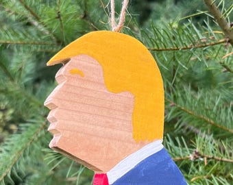 May include: A wooden Christmas ornament shaped like a person's head with yellow hair, a white collar, and a blue suit. The ornament is painted with red and white details.