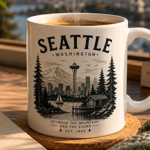 May include: A white ceramic mug featuring a black graphic illustration of the Seattle skyline, including the Space Needle, mountains, a sailboat, and a cabin. The text reads SEATTLE WASHINGTON, BETWEEN THE MOUNTAINS AND THE SOUND, and EST. 1869.