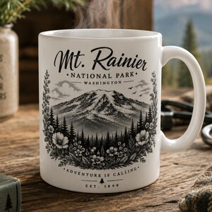 May include: A white ceramic mug featuring a black graphic illustration of Mt. Rainier National Park in Washington. The design includes a mountain peak, pine trees, and wildflowers, with the text Mt. Rainier National Park Washington and Adventure is Calling Est. 1899.