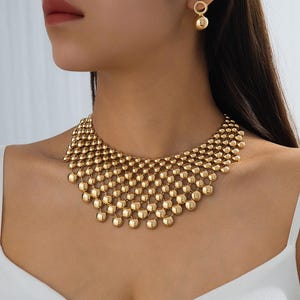 May include: A gold-toned statement necklace featuring a wide, multi-layered mesh design composed of numerous small, polished spherical beads linked together. The set includes matching drop earrings, each consisting of a small ring and a sphere.