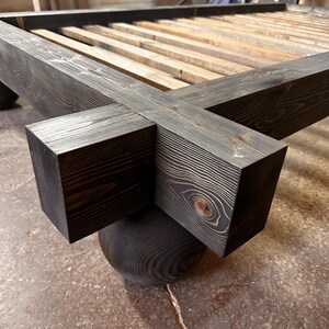 May include: Close-up of a dark stained wooden bed frame. The frame features large, square corner posts and a slatted base. The bed frame is supported by rounded, dark wooden feet. The wood grain is visible throughout the frame.