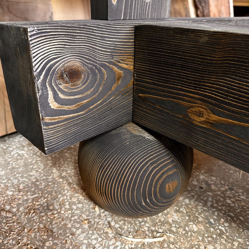 May include: Close-up of a wooden structure with a dark gray stain, showcasing the natural wood grain. The design features intersecting beams and a rounded base, highlighting the craftsmanship and texture of the wood.