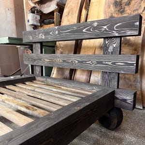May include: A handcrafted wooden bed frame with a dark gray stain. The headboard features two horizontal planks with visible wood grain. The bed frame has a slatted base and is supported by thick, square legs. The overall design is rustic and modern.