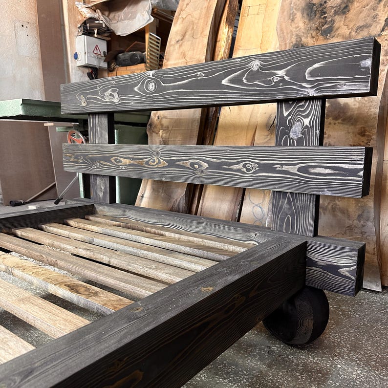May include: A handcrafted wooden bed frame with a dark gray stain. The headboard features two horizontal planks with visible wood grain. The bed frame has a slatted base and is supported by thick, square legs. The overall design is rustic and modern.