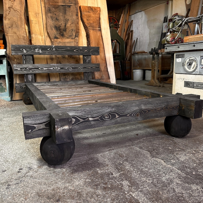 May include: A handcrafted, dark-stained wooden bed frame with a headboard and a slatted base. The bed frame is supported by four large, rounded wooden feet. The wood grain is visible throughout the frame, adding to its rustic charm. The bed frame measures approximately 78 inches long.