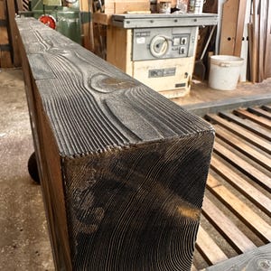 May include: Close-up of a large, rectangular, dark-stained wooden beam. The wood grain is prominent, with a textured, weathered appearance. The beam is likely for construction or decorative purposes.