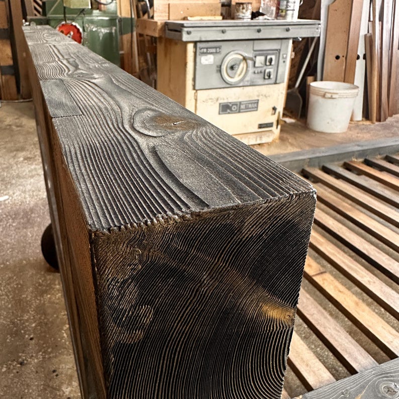 May include: Close-up of a large, rectangular, dark-stained wooden beam. The wood grain is prominent, with a textured, weathered appearance. The beam is likely for construction or decorative purposes.