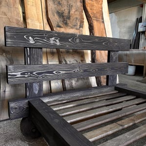May include: A handcrafted wooden bench with a dark gray finish. The bench features a slatted seat and a backrest with two horizontal planks. The wood grain is visible, adding texture. The design is rustic and suitable for indoor or outdoor use.