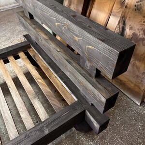 May include: A dark gray wooden bed frame with a slatted base. The headboard and footboard are constructed from thick, square timbers, showcasing the natural wood grain. The bed frame is designed with a modern, minimalist aesthetic.