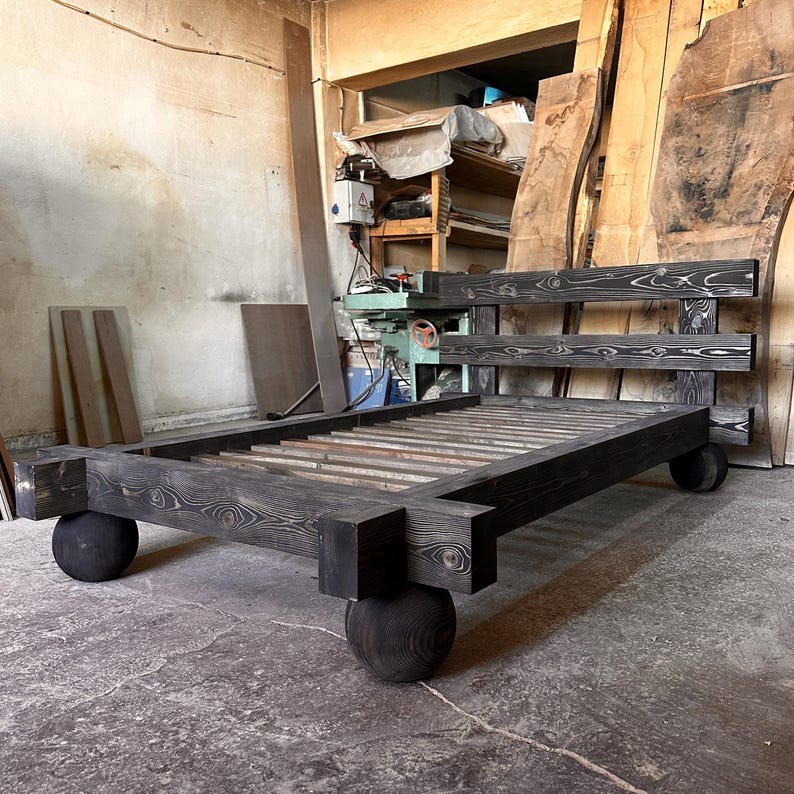 May include: A dark stained wooden bed frame with a headboard and footboard. The bed frame has a slatted base and rests on four large, round wooden feet. The wood grain is visible, and the bed appears to be handmade.