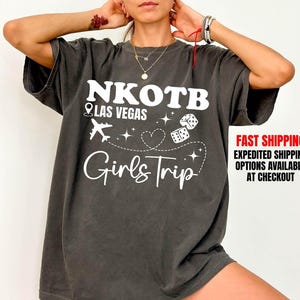 May include: Dark gray t-shirt with white text and graphics. The shirt reads "NKOTB Las Vegas Girls Trip" with an airplane, dice, and heart design. The shirt has short sleeves and a relaxed fit.