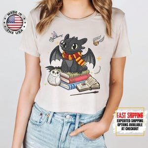 May include: A light beige t-shirt featuring a cartoon dragon wearing a scarf, sitting on books with an owl. The shirt has a "Made in the USA" logo on the upper left and "Fast Shipping" text on the lower right.