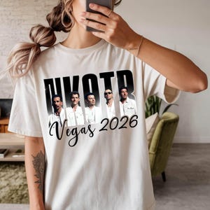 May include: Cream-colored t-shirt featuring the text "NKOTB" in bold black letters above a photo of five men. Below the photo, the words "Vegas 2026" are written in a cursive font. The shirt is a casual style.