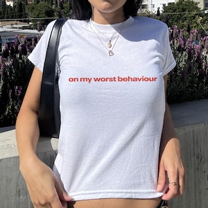Può includere: T-shirt bianco con la scritta "on my worst behaviour" stampata in rosso. La maglietta è a maniche corte e sembra essere realizzata in un materiale morbido. La modella indossa occhiali da sole e gioielli.