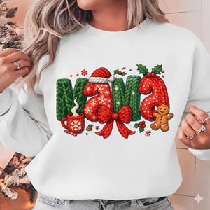 May include: White sweatshirt featuring the word "MAMA" in a festive design. The letters are green and red, with a Santa hat on the "M" and a bow on the "A". Includes a gingerbread man, snowflakes, and a mug of hot cocoa.