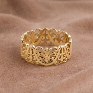 Könnte beinhalten: Ein goldfarbener Ring mit einem aufwendigen, durchbrochenen Design. Der Ring weist ein sich wiederholendes Muster aus floralen und scrollartigen Elementen auf, wodurch er zart und verziert wirkt. Der Ring ist vor einem weichen, neutralen Hintergrund platziert.