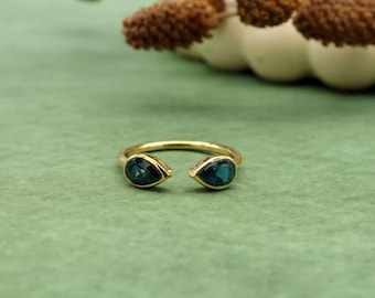 Op de afbeelding: Een goudkleurige ring met twee donkerblauwe, druppelvormige edelstenen. De ring heeft een open ontwerp, met de edelstenen aan de uiteinden van de band. De ring wordt gepresenteerd op een groene ondergrond.