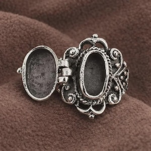Op de afbeelding: Een zilverkleurige locket ring met een ovaal compartiment dat opengaat. De ring heeft een sierlijk, filigraan ontwerp met decoratieve krullen en een kroonachtig detail. De locket ligt op een bruin oppervlak.