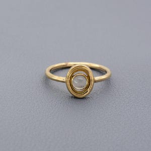 Pode incluir: Um anel dourado com um engaste oval. O engaste apresenta uma pedra central transparente rodeada por um design em espiral. A banda do anel é fina e simples. O anel é exibido numa superfície cinzenta.