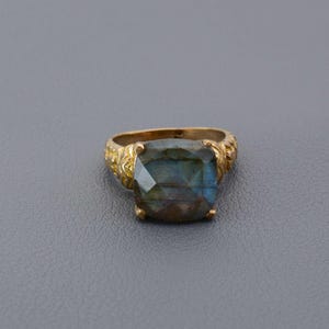 Könnte beinhalten: Ein goldfarbener Ring mit einem großen, quadratisch geschliffenen Labradorit-Edelstein. Der Stein zeigt schillernde Farben von Blau, Grün und Braun. Das Ringband hat ein verziertes, strukturiertes Design.