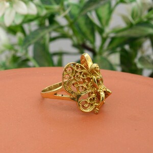 May include: A gold-colored ring featuring a detailed fairy design. The fairy has outstretched wings and is positioned on a ring band. The ring is set against a terracotta-colored surface with a blurred green background.