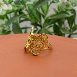 May include: A gold-colored ring featuring a detailed filigree design with a central figure. The ring has an ornate, openwork pattern and a polished finish. The ring is set against a blurred green and brown background.
