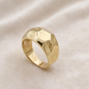 May include: A gold-colored ring with a faceted design. The ring has a wide band and a geometric pattern across the top, reflecting light. The ring is set against a soft, off-white background.