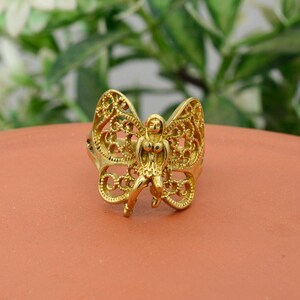 May include: A gold-colored ring featuring a butterfly design with a central figure. The butterfly wings have an openwork filigree pattern. The ring is set against a terracotta-colored surface.
