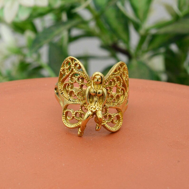 May include: A gold-colored ring featuring a butterfly design with a central figure. The butterfly wings have an openwork filigree pattern. The ring is set against a terracotta-colored surface.