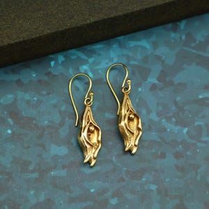 以下が含まれることがあります： 金色のイヤリングのペア。各イヤリングは、湾曲したフックが付いた細長い花の形をしています。イヤリングは、青と緑のテクスチャード加工された背景に配置されています。イヤリングの長さは約3cmです。