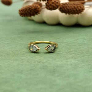 Può includere: Un anello aperto color oro con due pietre preziose a forma di pera, verde chiaro. L'anello è posto su uno sfondo verde, con elementi decorativi sfocati sullo sfondo.