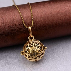 Può includere: Una collana con pendente dorato. Il pendente è una sfera in filigrana con intricati motivi floreali e una piccola gemma. La catena della collana è una delicata catena a serpente dorata. Lo sfondo è una superficie marrone strutturata.