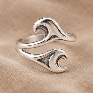 Könnte beinhalten: Ein offener Ring aus poliertem Silber mit zwei symmetrischen, geschwungenen, wellenartigen Motiven, die einander zugewandt sind. Die Metalloberfläche hat ein glattes, reflektierendes Finish mit dunklen, vertieften Bereichen, die das Design betonen.