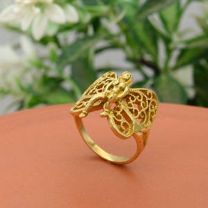 May include: A gold-colored ring featuring a detailed angel design. The ring has an openwork wing design and a small figure of an angel. The ring is set against a blurred background of green foliage and a terracotta surface.