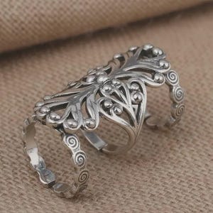 Può includere: Un anello argentato a più dita con un design a filigrana ornato. L'anello presenta un motivo floreale centrale con dettagli a volute e piccoli accenti rotondi. L'anello è progettato per coprire due dita, con fasce regolabili.