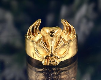 May include: A gold-colored ring featuring a detailed cobra design with outstretched wings. The ring has a wide band and intricate detailing, reflecting light. The ring's design is centered around the cobra's head, with wings on either side.