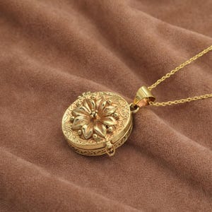 May include: A gold locket necklace featuring an oval pendant with a floral design. The pendant has an embossed flower and detailed patterns. The necklace has a fine chain and a clasp. The locket is displayed on a brown surface.