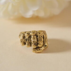 May include: A gold-coloured ring with a clenched fist design and skull details. The ring has a detailed, textured surface. The design incorporates multiple skulls. The ring is set against a soft, neutral background.