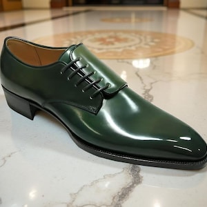May include: A single, glossy, dark green leather dress shoe with black laces. The shoe has a square toe and a low heel. The shoe is sitting on a marble surface.