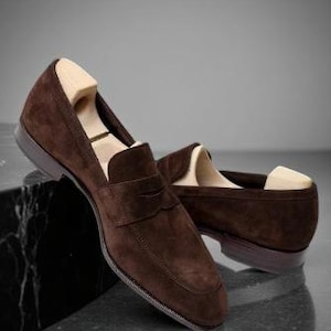 May include: A pair of brown suede penny loafers with wooden shoe trees inside. The shoes are displayed on a dark, reflective surface, showcasing their classic design and rich color. The loafers have a rounded toe and a low heel.