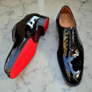 May include: Pair of glossy black dress shoes with red soles. The shoes have a classic lace-up design and a high-shine finish. The interior of the shoe has a logo. The shoes are displayed on a marble surface.