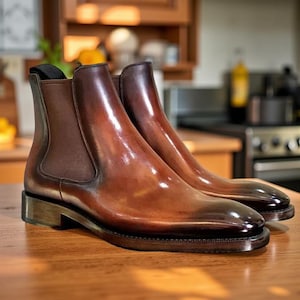 Könnte beinhalten: Ein Paar braune Chelsea-Stiefel aus Leder mit glänzender Oberfläche. Die Stiefel haben ein klassisches Design mit elastischen Seiteneinsätzen und einem niedrigen Absatz. Das Leder hat einen Farbverlauf, der von einem helleren Braun zu einem dunkleren Farbton an der Spitze übergeht.