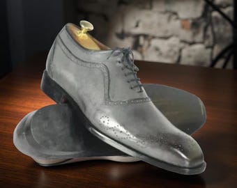 May include: A pair of gray leather dress shoes with black soles and laces. The shoes have decorative perforations and a wooden shoe tree inside. The shoes are displayed on a wooden surface.
