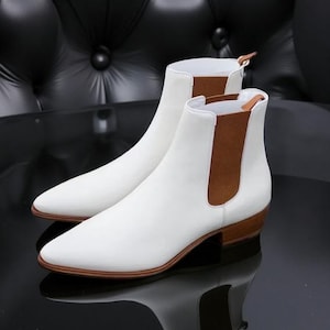 Peut inclure: Une paire de bottines Chelsea en cuir blanc avec un bout pointu et des empiècements latéraux élastiques marron. Les bottines ont un petit talon empilé et une semelle marron. Les bottines sont présentées sur une surface noire réfléchissante.