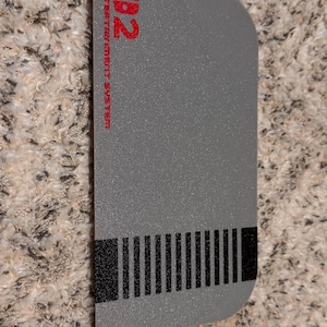 Puede incluir: Una pegatina rectangular gris con un diseño de videojuego retro. La pegatina presenta el texto "02 ENTERTAINMENT SYSTEM" en rojo, junto con un diseño en negro y gris que recuerda a un cartucho de juego clásico. La pegatina está sobre una superficie texturizada.