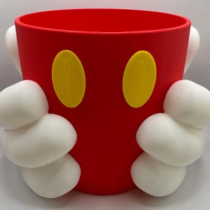 Puede incluir: Un vaso de plástico rojo con dos formas ovaladas amarillas y asas blancas y redondeadas. El vaso está diseñado para parecerse a un personaje de dibujos animados.