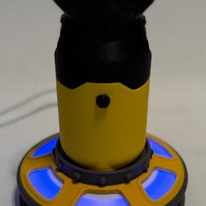 May include: A device with a black top and base, a yellow midsection, and a circular base with blue lights. The top has a red accent. The device appears to be made of plastic or a similar material.