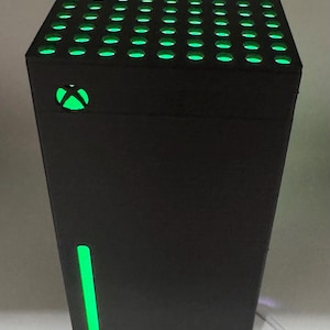 Pode incluir: Console de jogos preta com detalhes verdes iluminados. A parte superior tem numerosos orifícios pequenos e espaçados uniformemente com luz verde. O logotipo da Xbox é visível, também iluminado em verde. Há uma faixa de luz verde vertical.